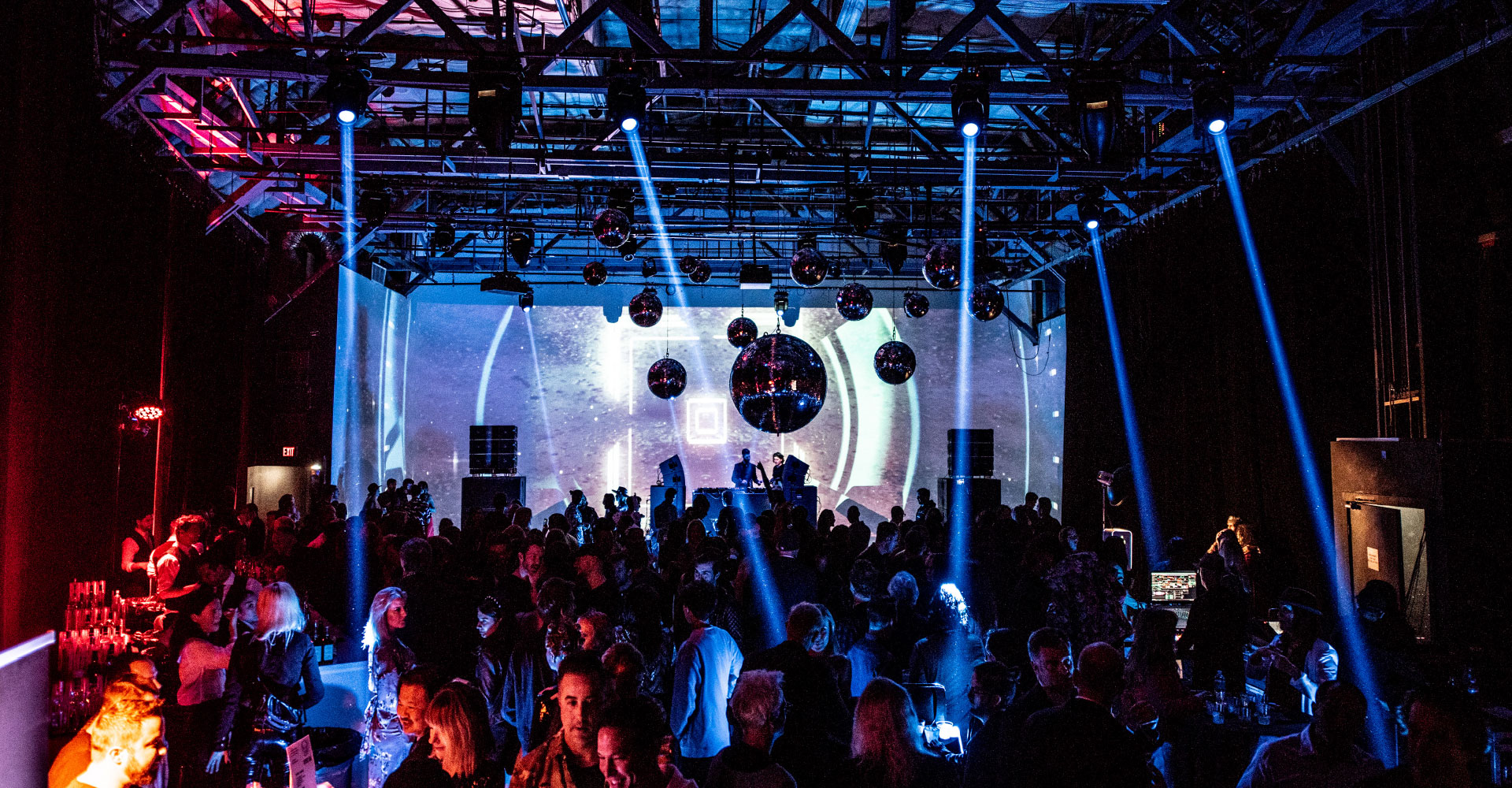 Wide view of the venue's dance floor and stage, crossing beams of blue and pink light sweeping over a large crowd gathered before a lit archway backdrop.