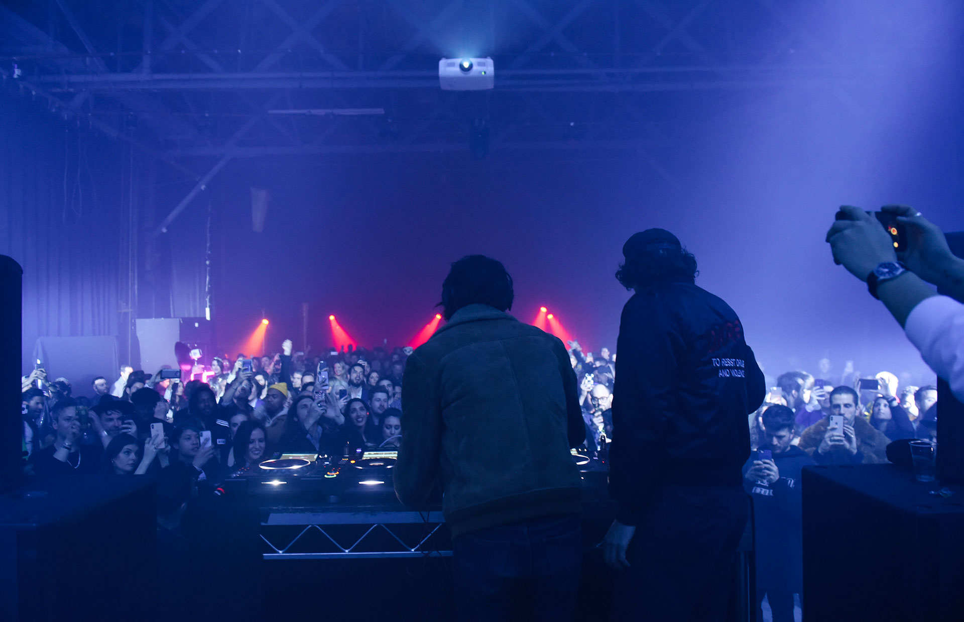 Two DJs seen from behind at the turntables, silhouetted against a haze of blue light as a packed crowd raises phones toward the stage, a projector suspended overhead.