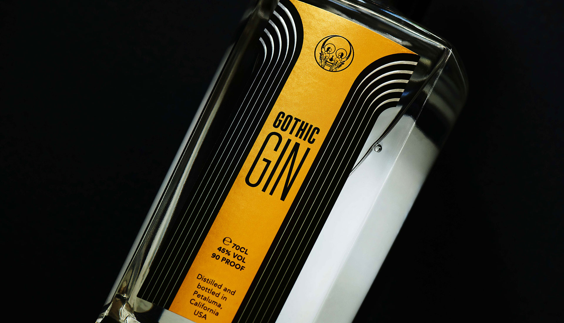 Close-up of the Gothic Gin bottle, its gold gothic-arch label and skull emblem catching dramatic light against a black background.