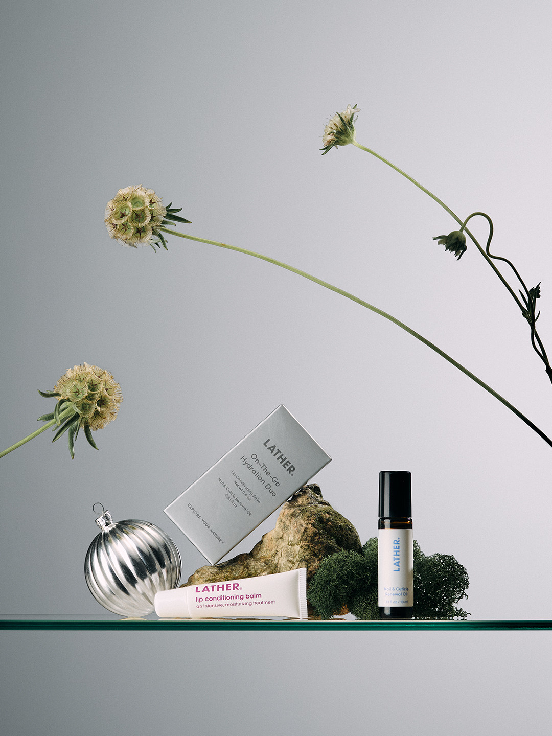 LATHER's On-The-Go Hydration Duo box, lip conditioning balm, and renewal oil styled with a silver ornament, a small rock, and dried scabiosa flower stems.