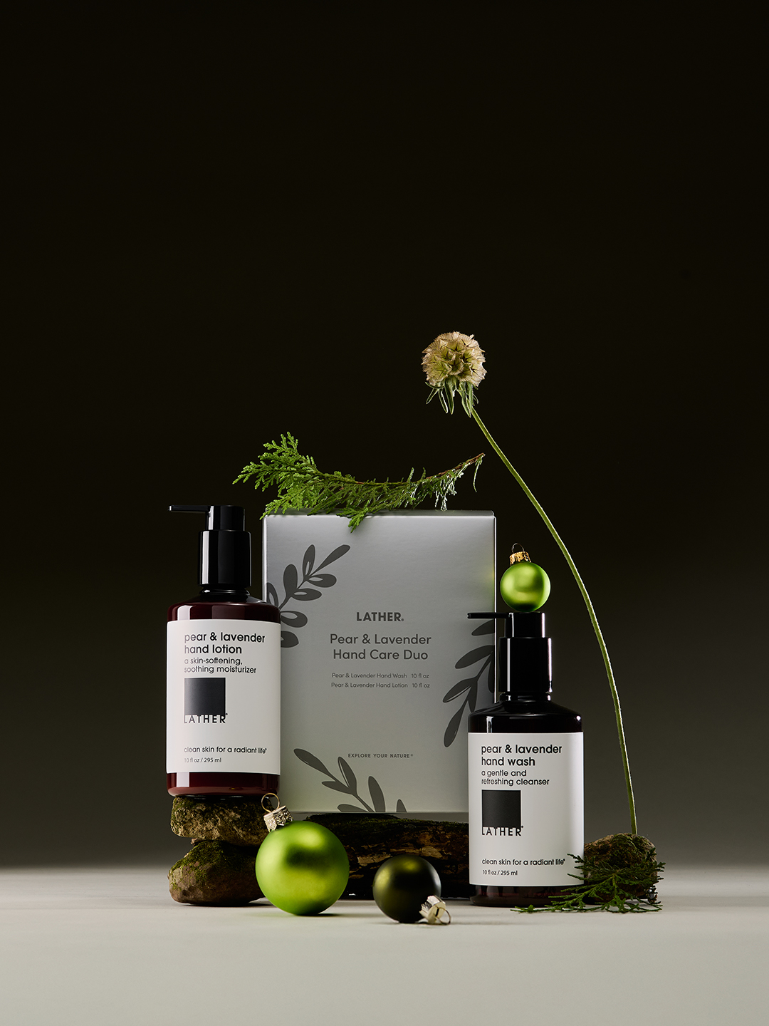 LATHER's Pear & Lavender hand lotion and hand wash bottles arranged with the boxed Hand Care Duo, a cypress branch, a dried flower, and green ornaments against a near-black background.