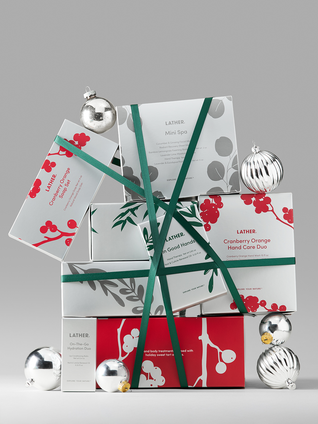 A tower of LATHER holiday gift boxes — including the Cranberry Orange Hand Care Duo, In Good Hands, Mini Spa, and On-The-Go Hydration Duo — bound with green ribbon and surrounded by silver ornaments.