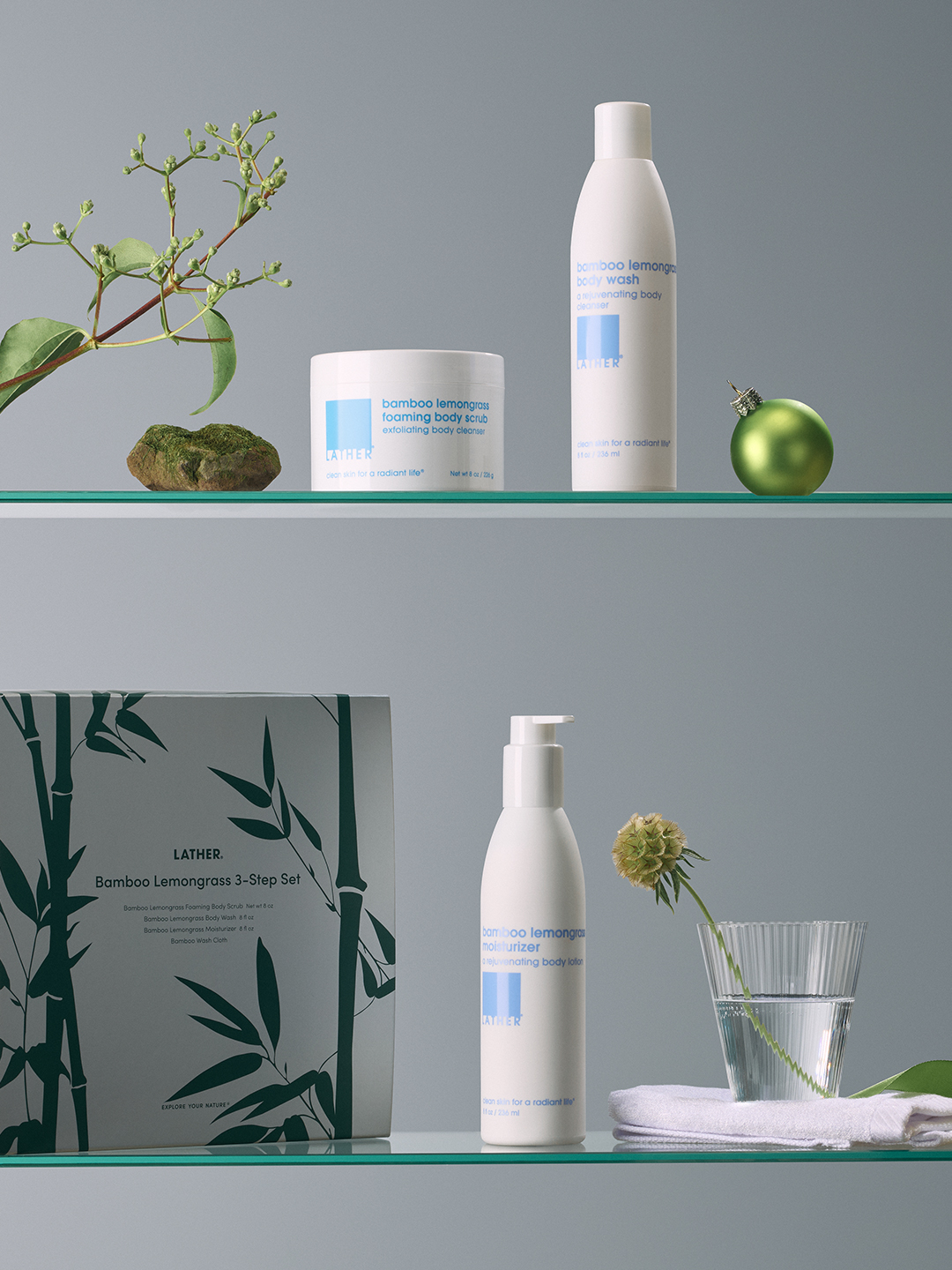 LATHER Bamboo Lemongrass foaming body scrub, body wash, moisturizer, and boxed 3-Step Set arranged on two glass shelves with a eucalyptus sprig, a green ornament, and a sprig in a glass of water.