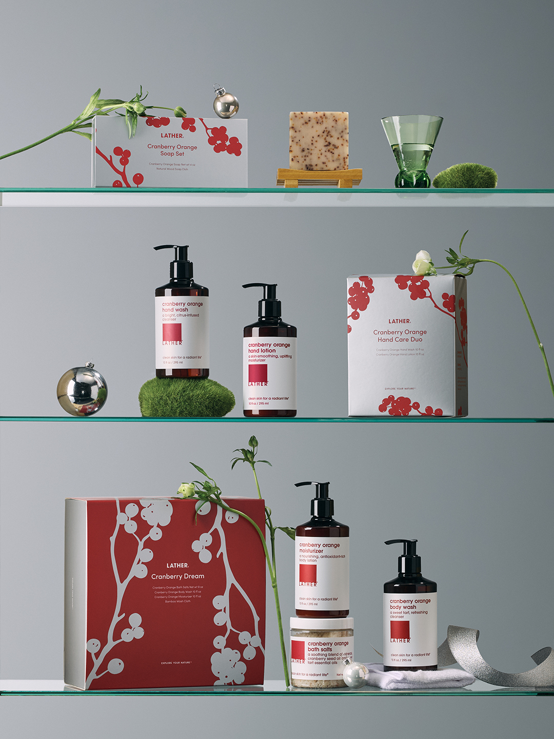 LATHER Cranberry Orange gift sets — a soap set, hand wash and lotion duo, hand care duo box, and the red-and-white Cranberry Dream box — arranged across three glass shelves with cranberry sprigs and a silver ornament.