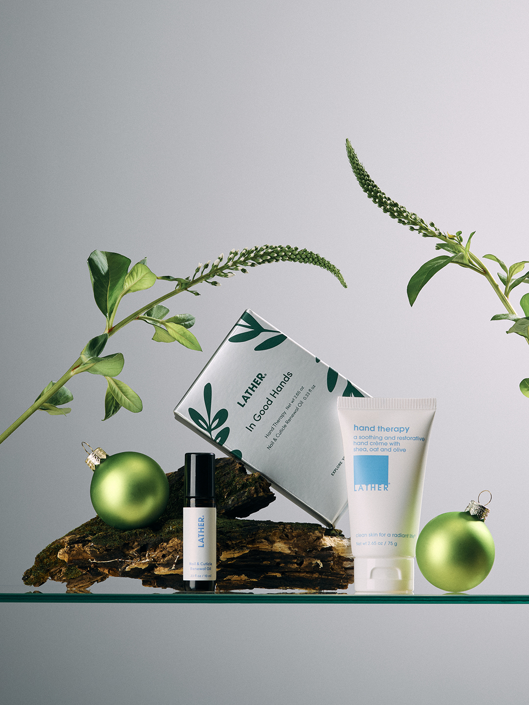 The LATHER In Good Hands gift box, hand therapy cream, and nail and cuticle renewal oil arranged on a piece of bark with fresh green foliage and ornaments.