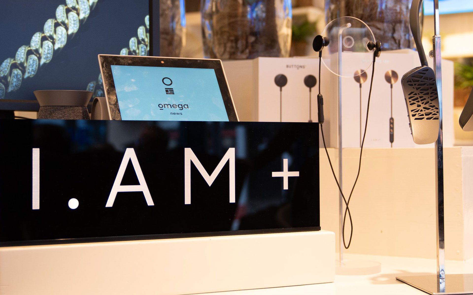 Retail display of I.AM+ products: a black sign reading 'I.AM+' beside a tablet showing the Omega news app interface, with Buttons earphones displayed on stands behind it.