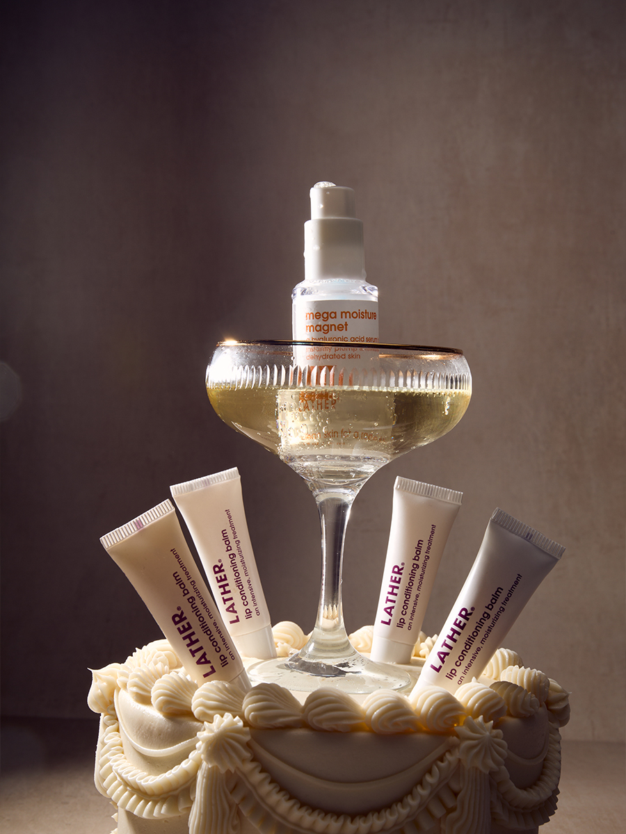 Lather lip conditioning balm tubes and a serum bottle arranged around a champagne coupe atop a frosted tiered cake.