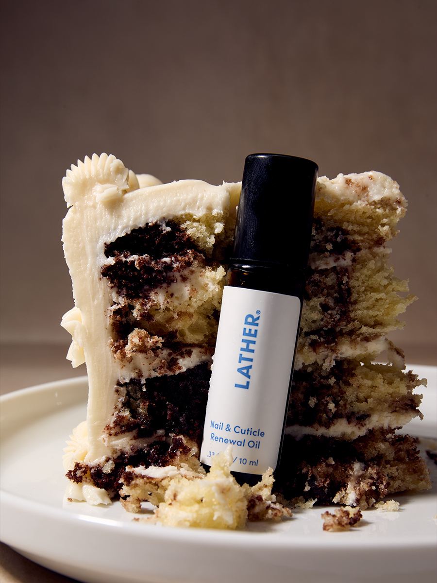 A rollerball bottle of Lather nail & cuticle renewal oil leaning against a slice of frosted layer cake on a white plate.