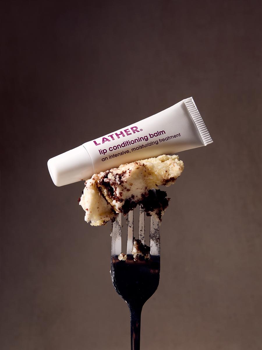 A tube of Lather lip conditioning balm balanced on a fork holding a bite of frosted layer cake.