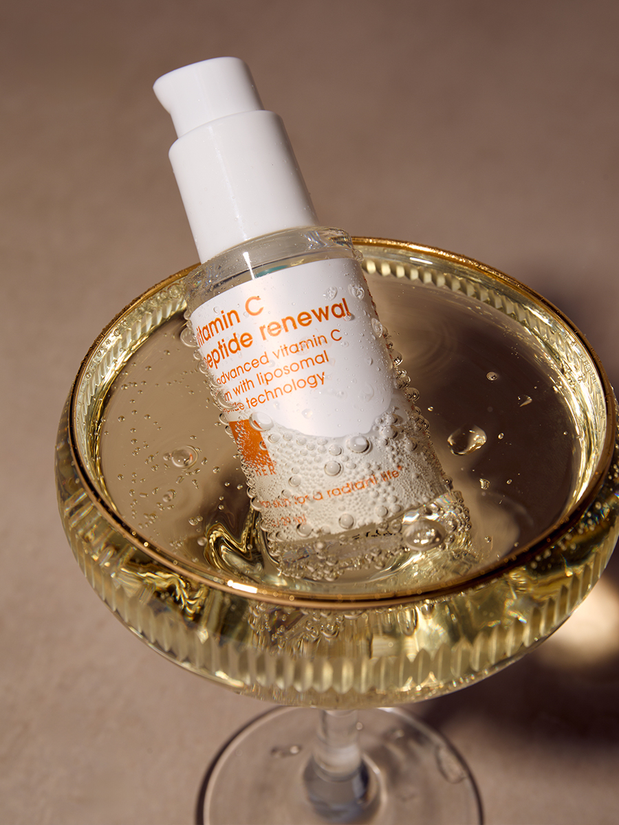 A bottle of Lather vitamin C peptide renewal serum resting in a coupe glass of champagne, warm golden light.