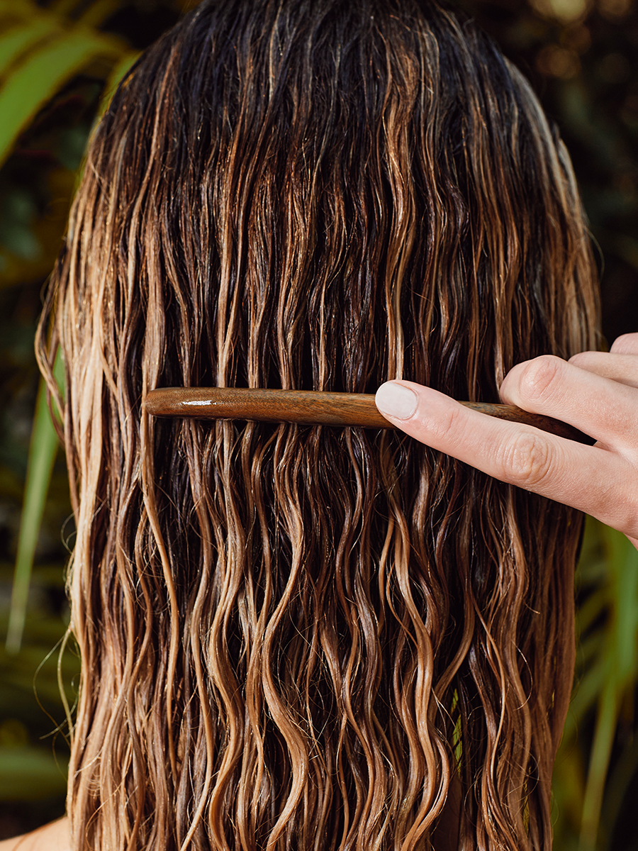 Rear view of a model's wet, highlighted waves being combed through with a wooden comb, tropical foliage behind.
