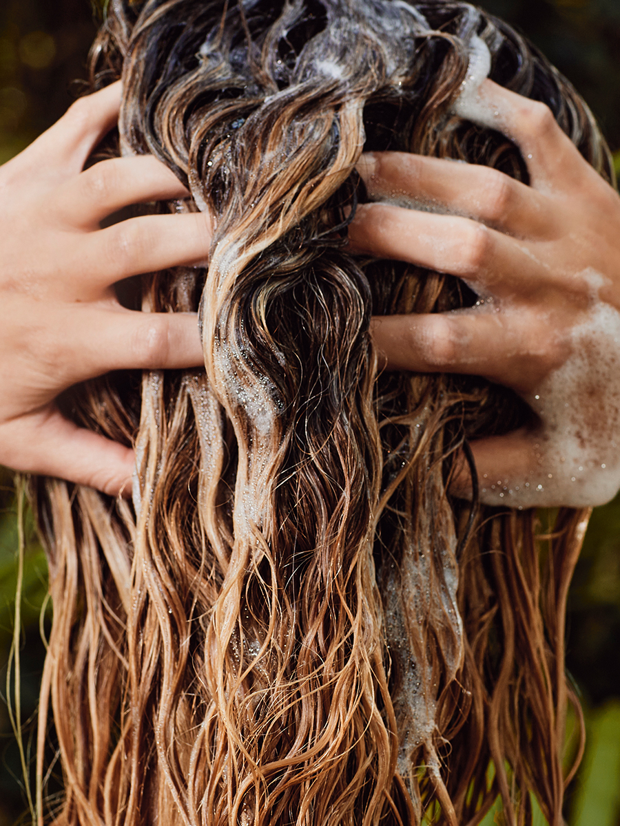 Close-up of hands massaging foaming shampoo into wet, wavy hair from above.