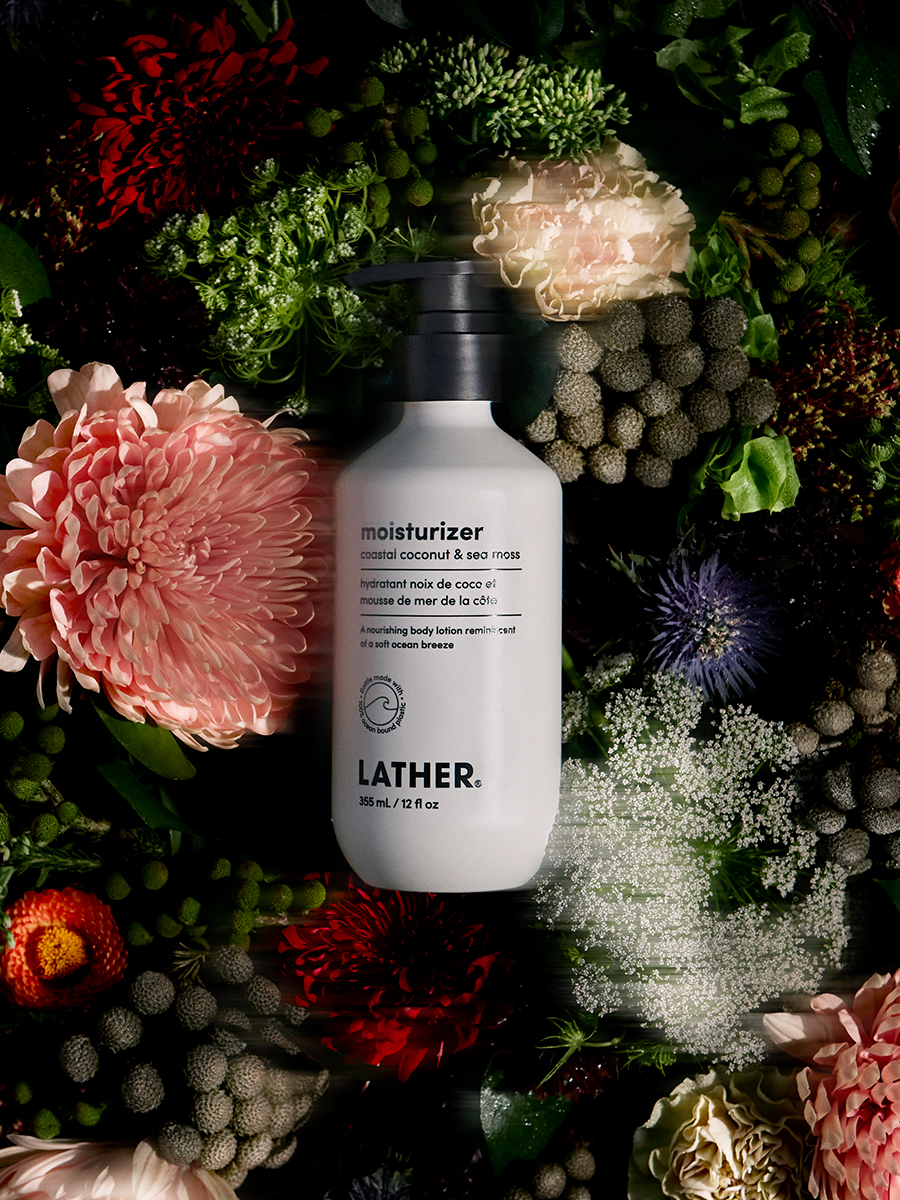 A bottle of Lather coastal coconut & sea moss moisturizer laid amid a dark, moody arrangement of pink and cream chrysanthemums and greenery.