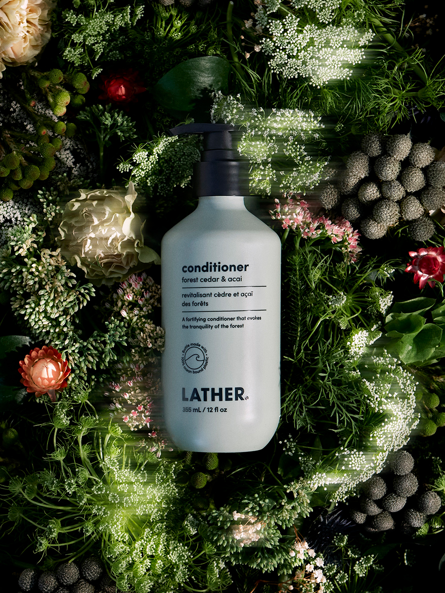 A bottle of Lather forest cedar & acai conditioner nestled in a dark bed of ferns, thistle, and white florals.