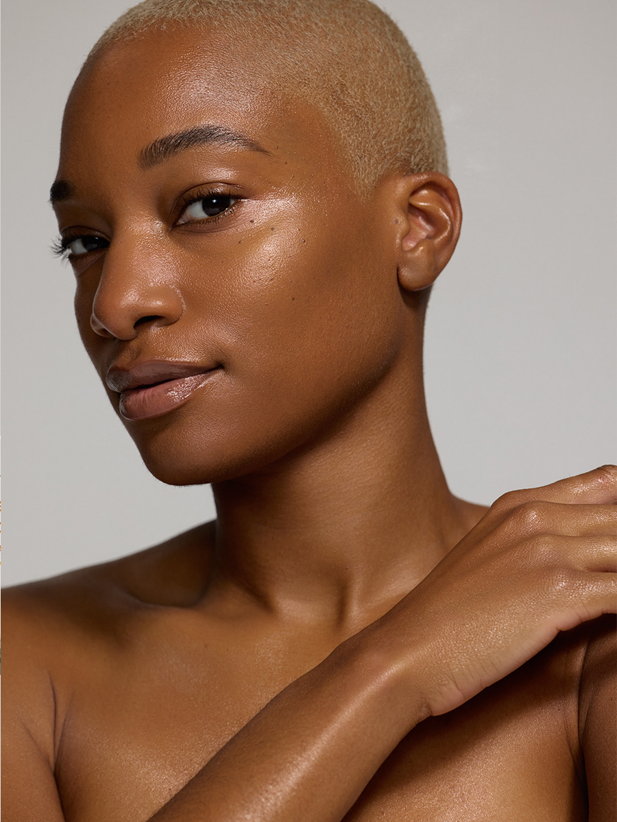 Close-up beauty portrait of a model with a bleached buzz cut and dewy skin, hand resting on her shoulder, softly lit against a warm grey backdrop.