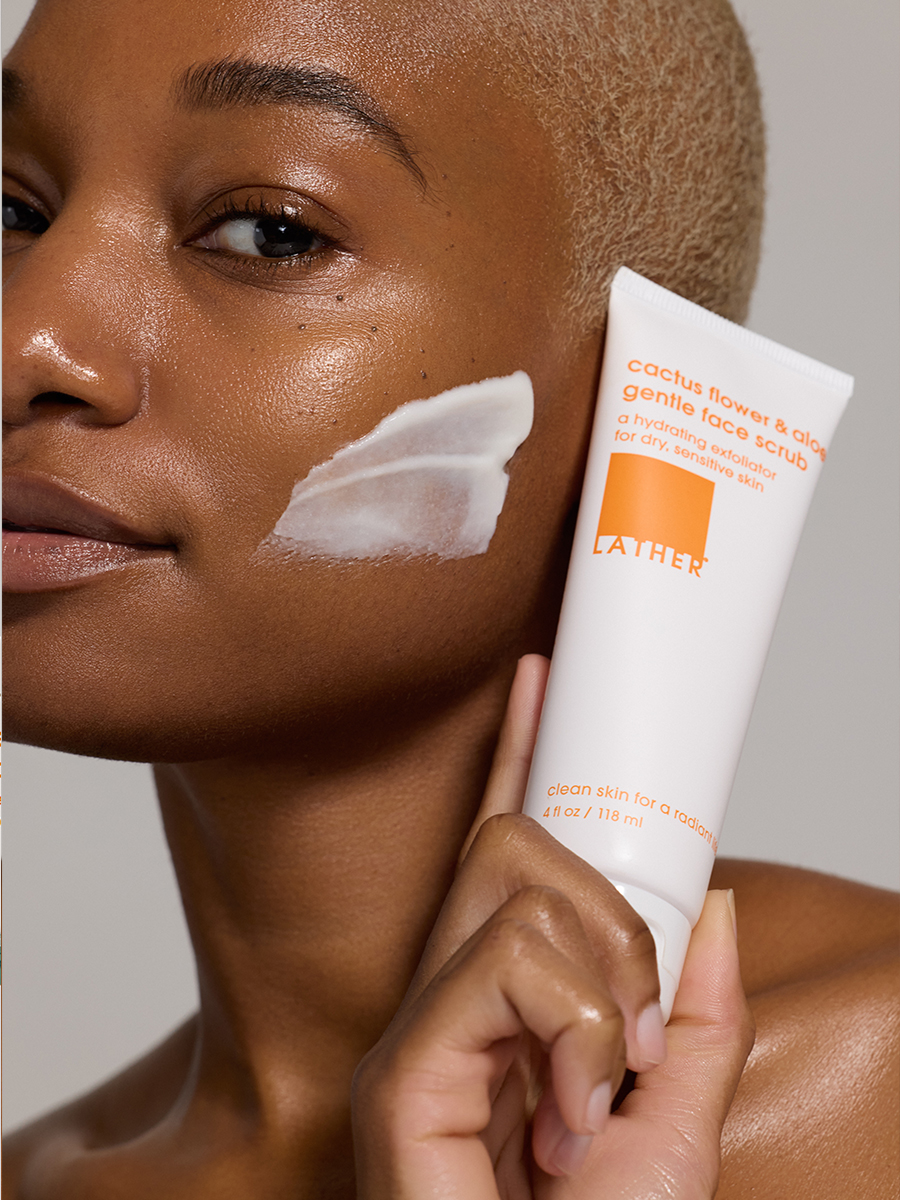 A model holds a tube of Lather cactus flower & aloe gentle face scrub beside her cheek, a swipe of the scrub applied to her skin.