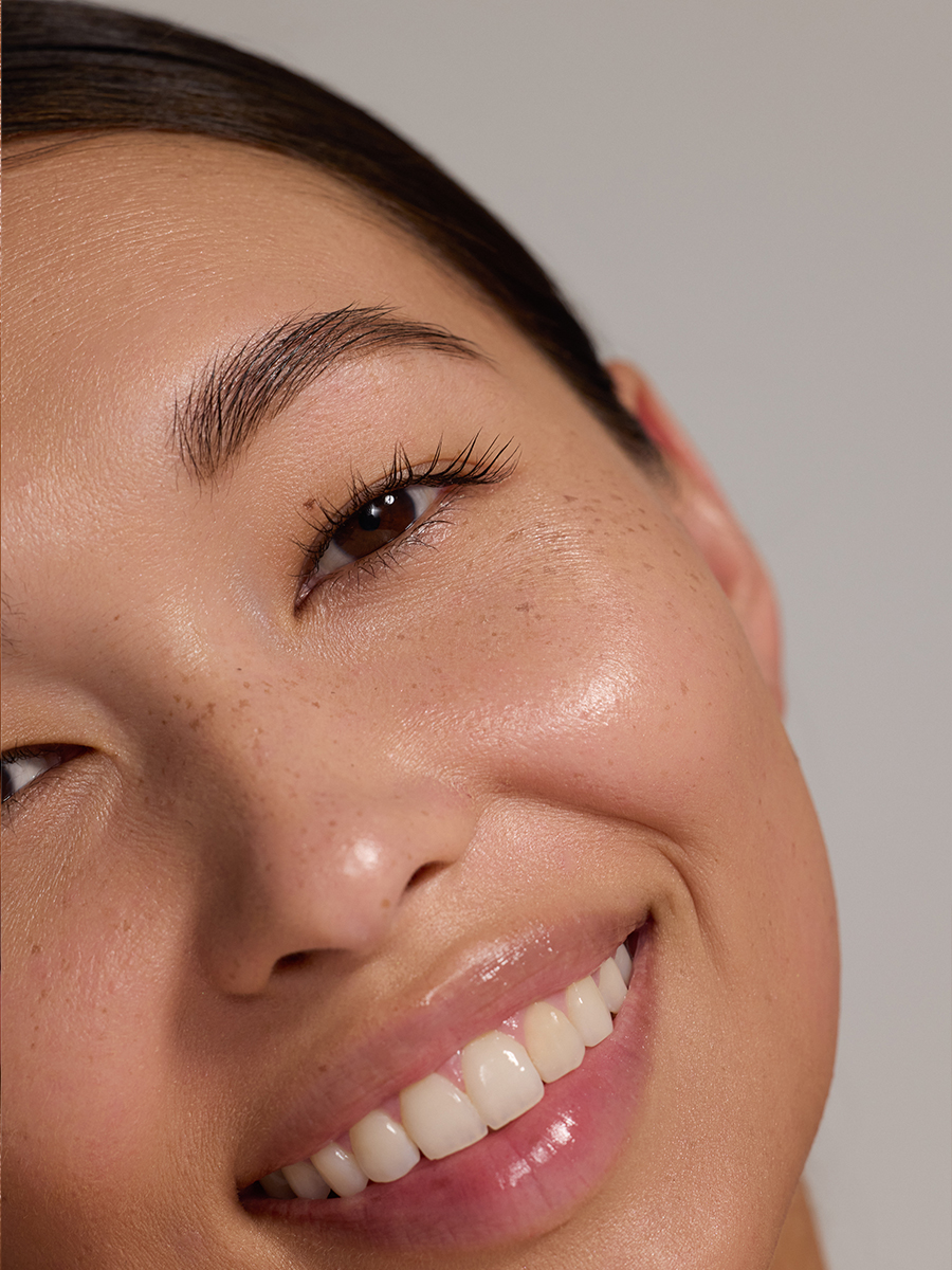 Close-up of a model's face turned upward, laughing, with freckles and glossy skin visible under warm light.