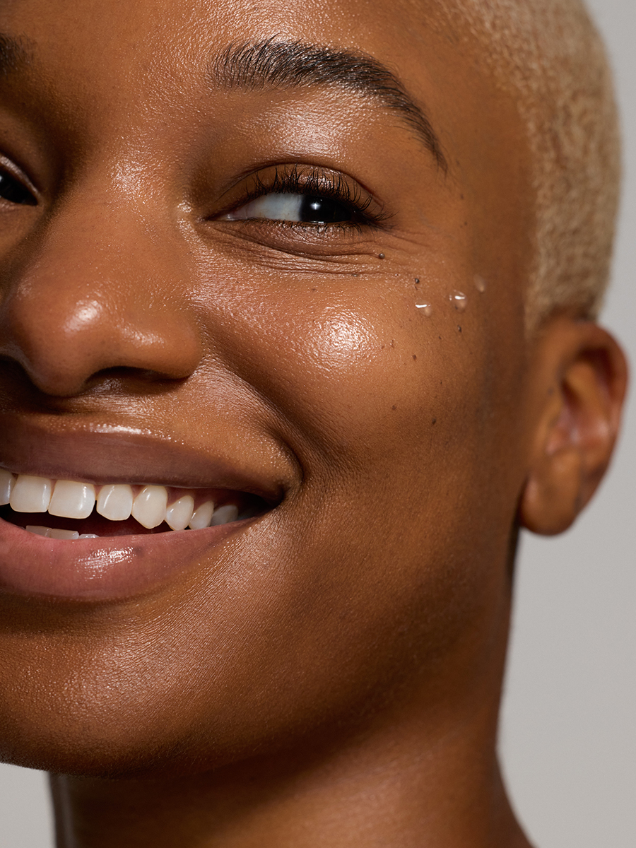 Close-up of a smiling model's face and eye, a drop of serum catching light near her cheekbone, bleached buzz cut visible.