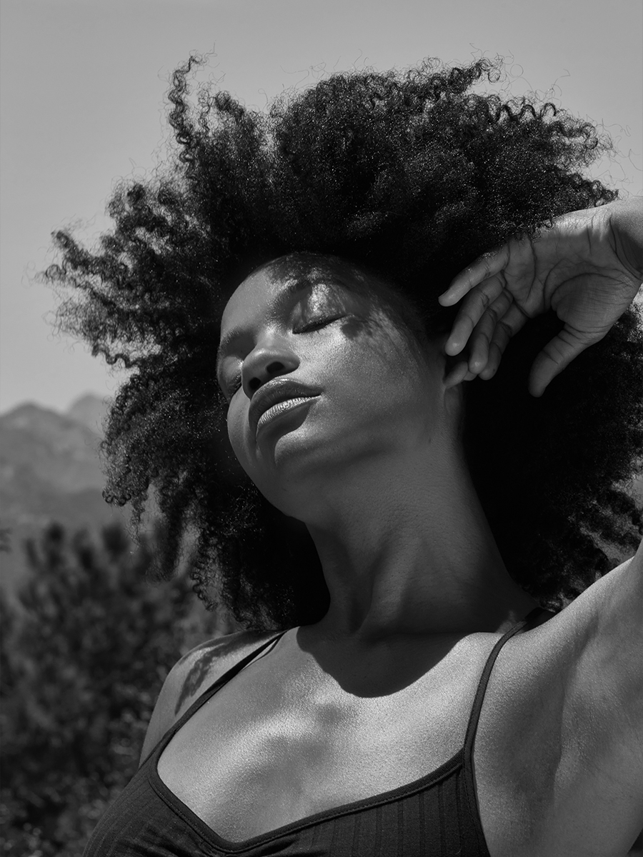 Black-and-white portrait of a model with windblown curly hair, eyes closed, mountains visible in the background.