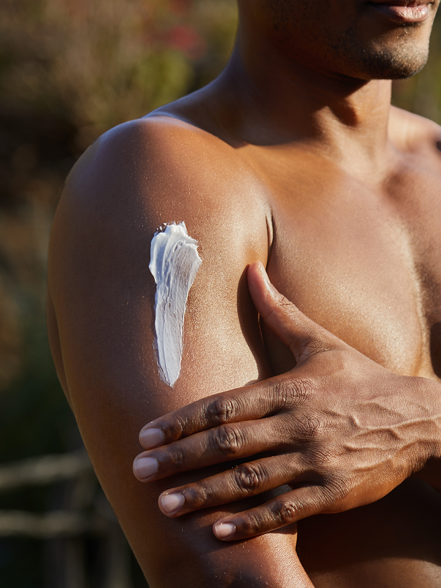 Black-and-white close-up of a man's shoulder and chest with a swipe of cream being rubbed in, outdoors.