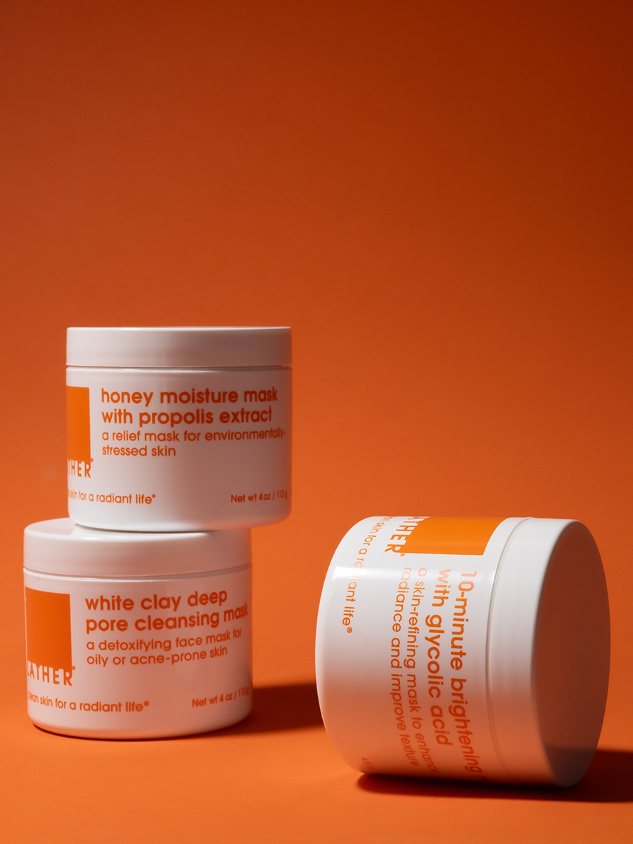 Three stacked Lather jars — honey moisture mask, white clay pore cleansing mask, and glycolic acid brightening mask — on a bright orange background.
