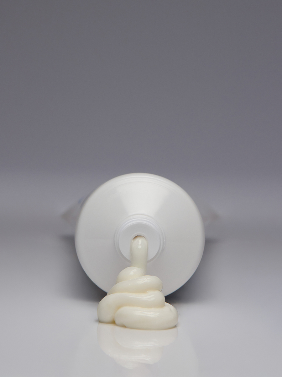 Extreme close-up of a white Lather tube squeezed to dispense a dollop of cream, shot head-on against a grey background.
