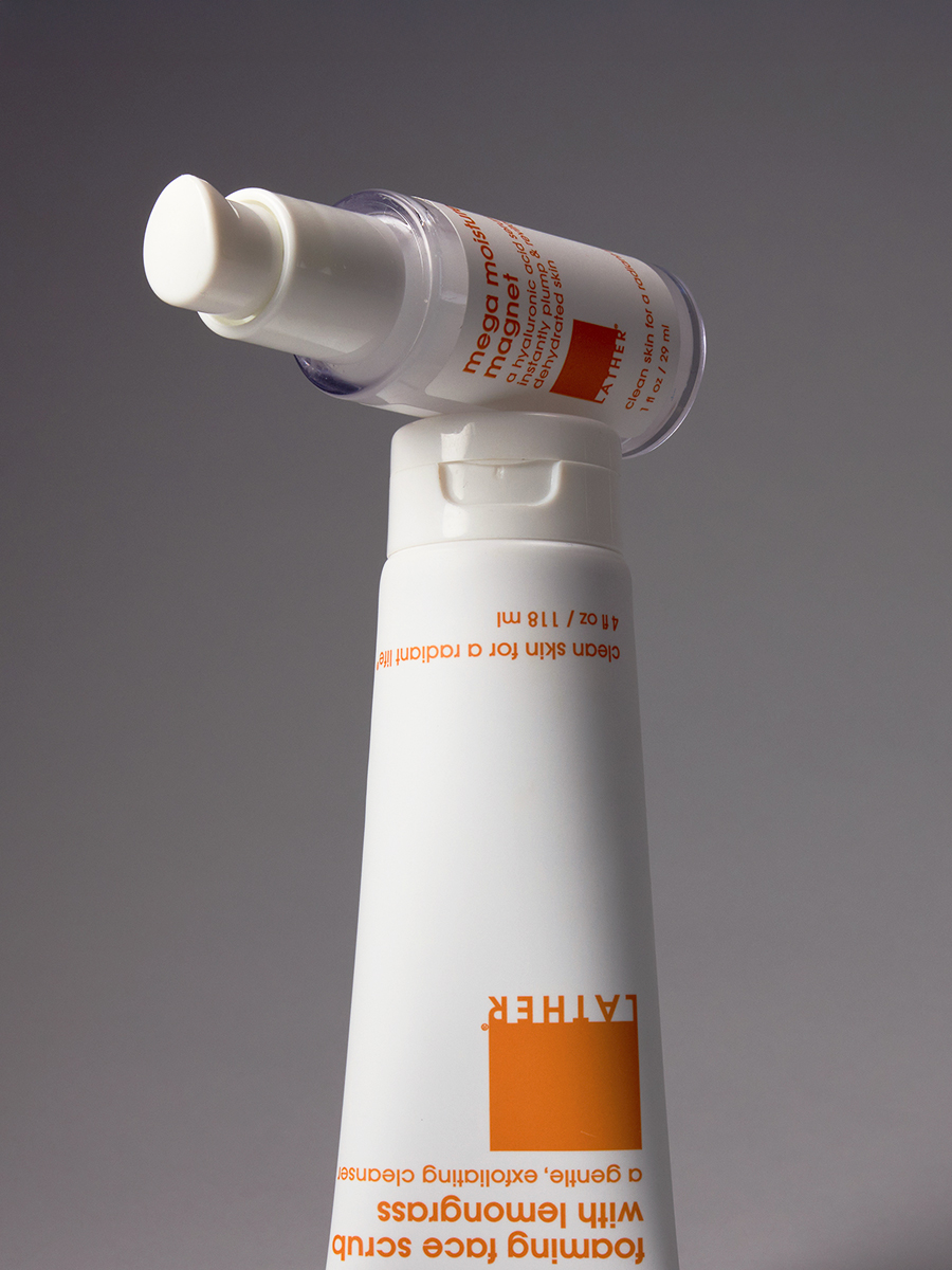 An upside-down bottle of Lather mega moisture magnet hyaluronic serum balanced atop an inverted tube of bamboo lemongrass foaming face scrub.