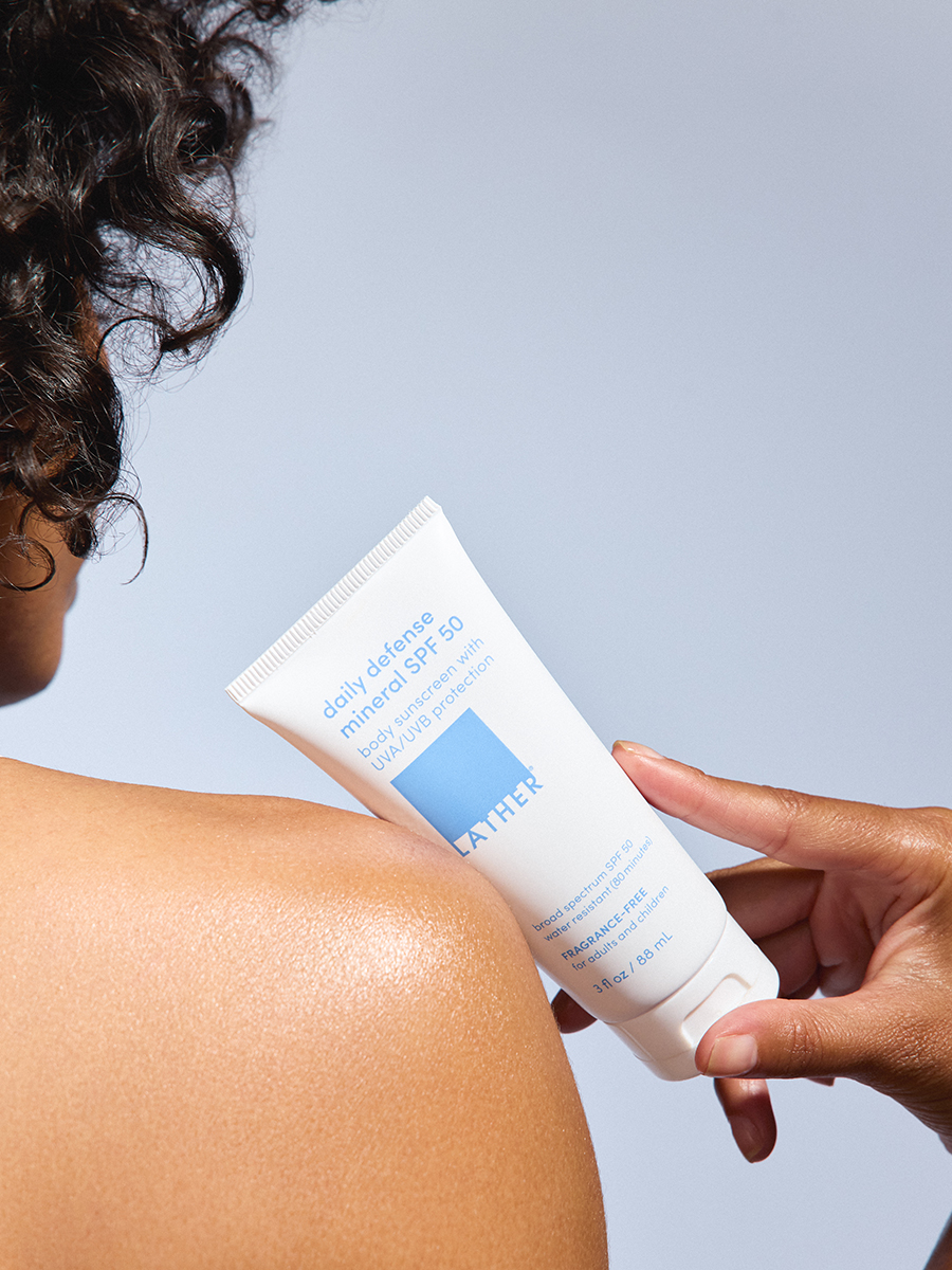 Close-up of a hand pressing a tube of Lather daily defense mineral SPF 50 against a sunlit shoulder.