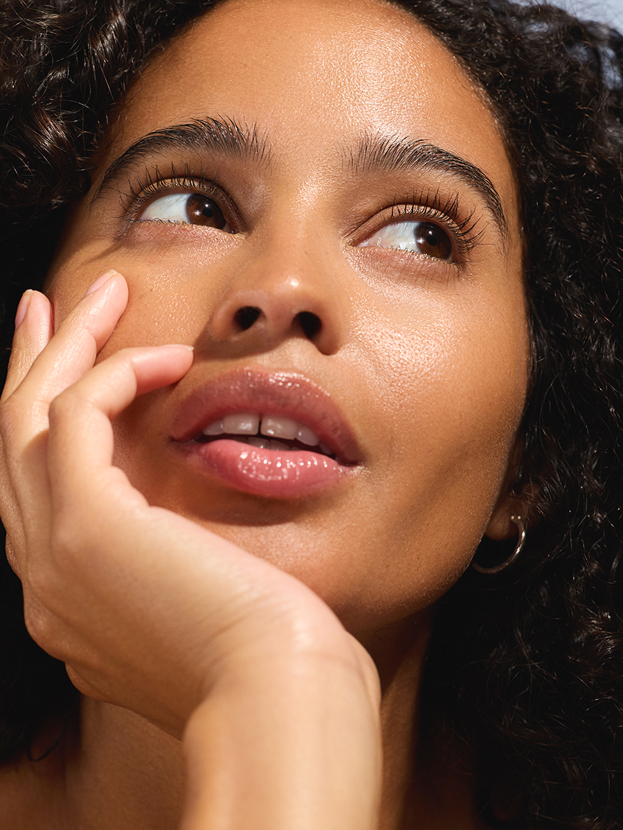 Close-up of a model's face tilted upward in sunlight, hand resting against her cheek, eyes gazing up.