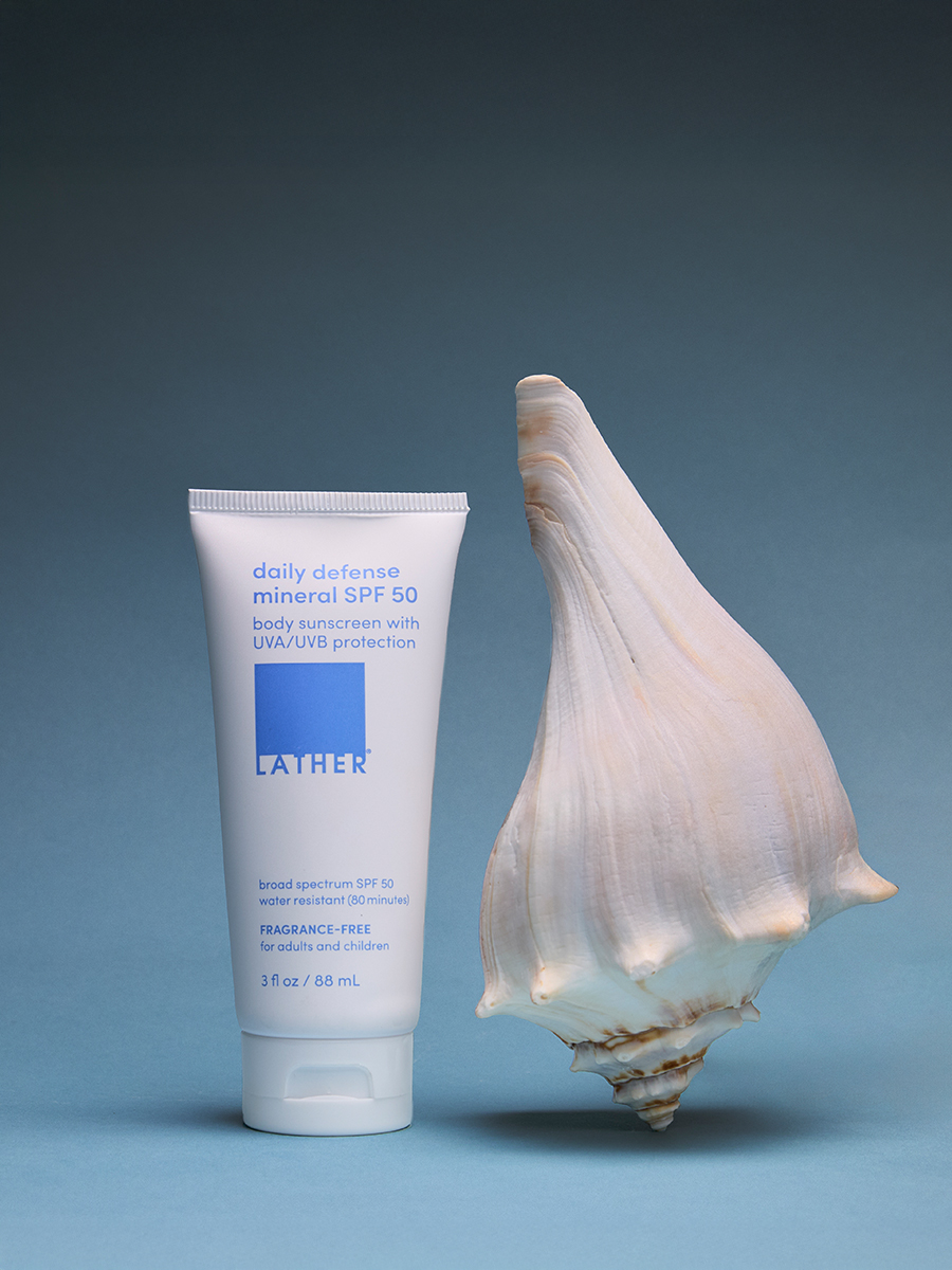 A tube of Lather daily defense mineral SPF 50 standing beside a large spiral seashell on a blue background.