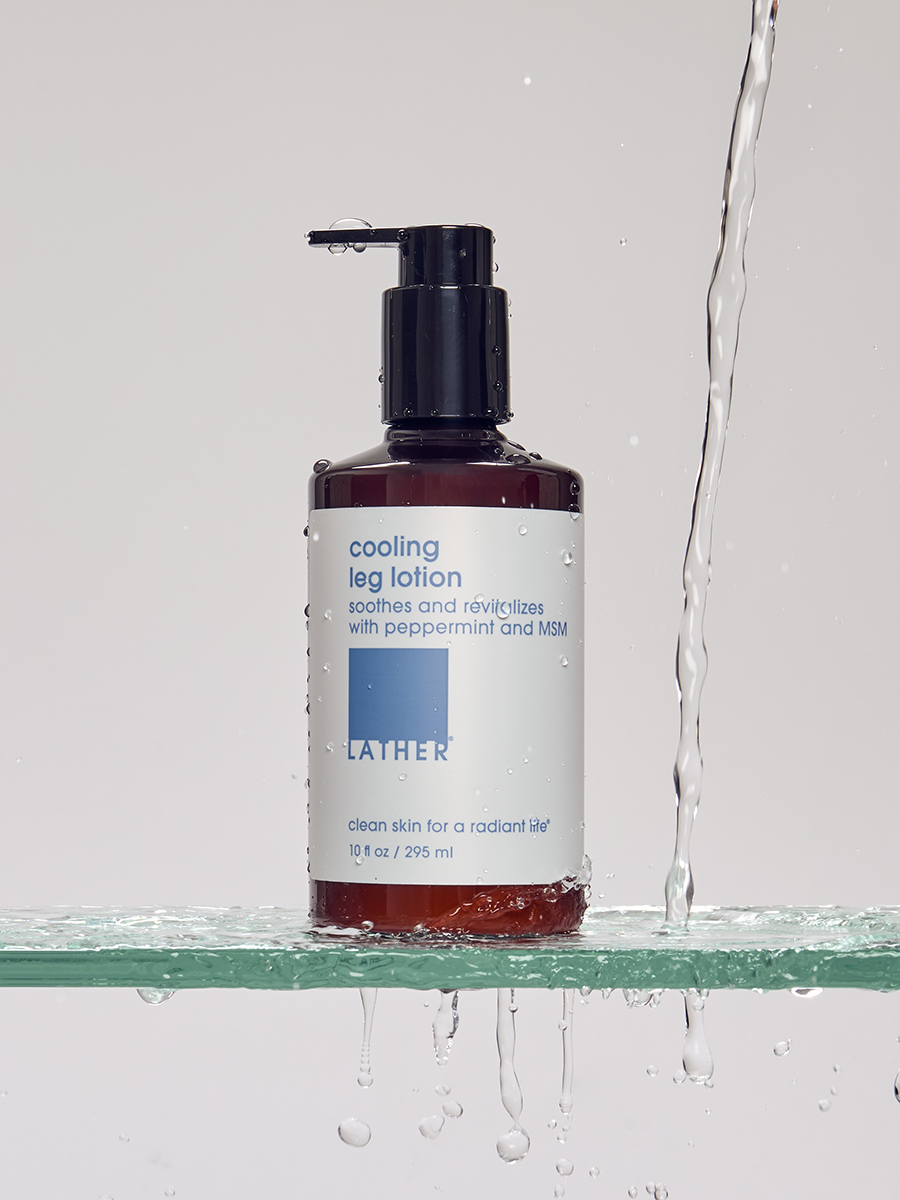 A bottle of Lather cooling leg lotion balanced on a glass shelf as water streams and splashes around it.