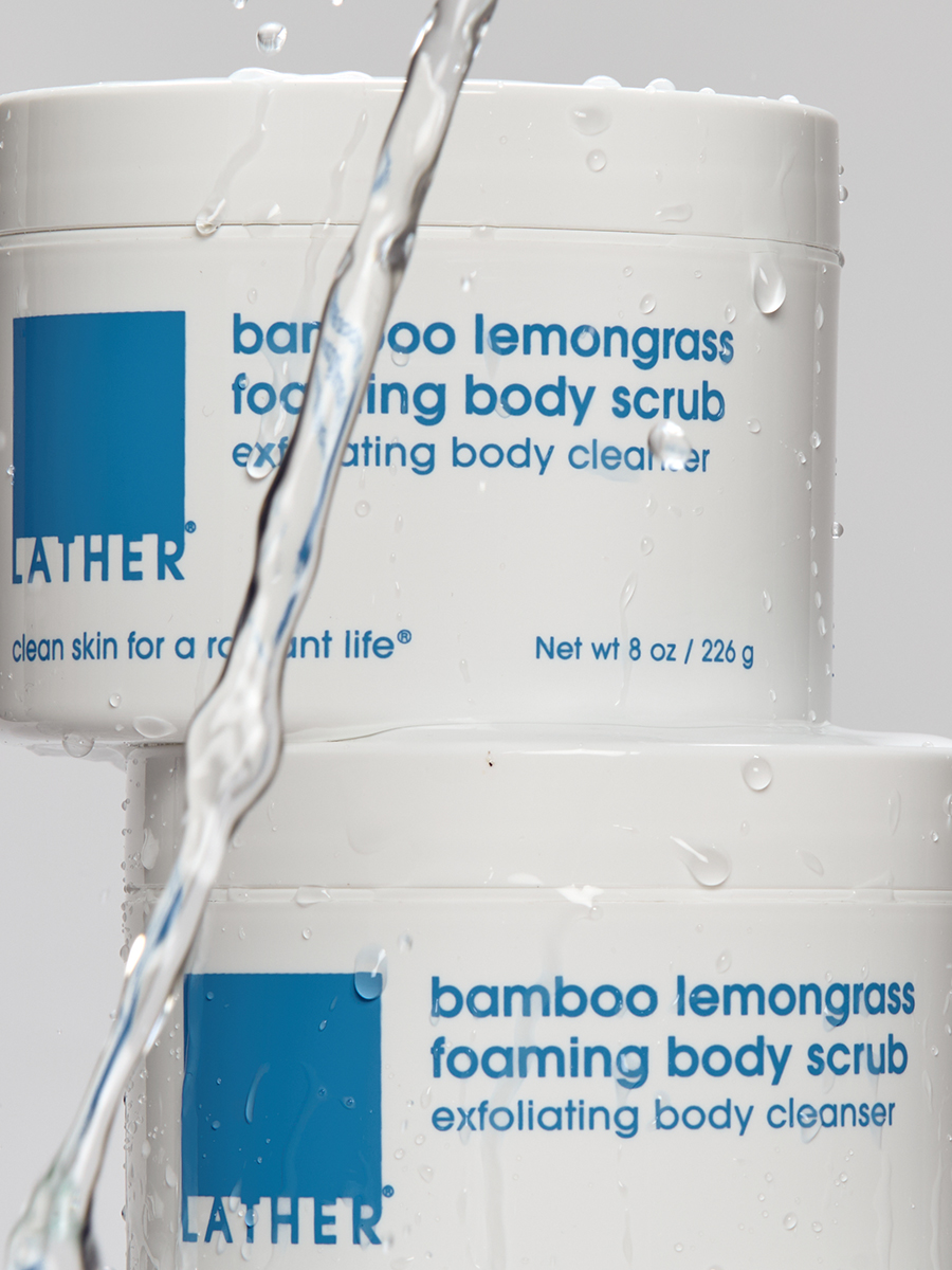Two bottles of Lather bamboo lemongrass foaming body scrub stacked as water pours down between them.