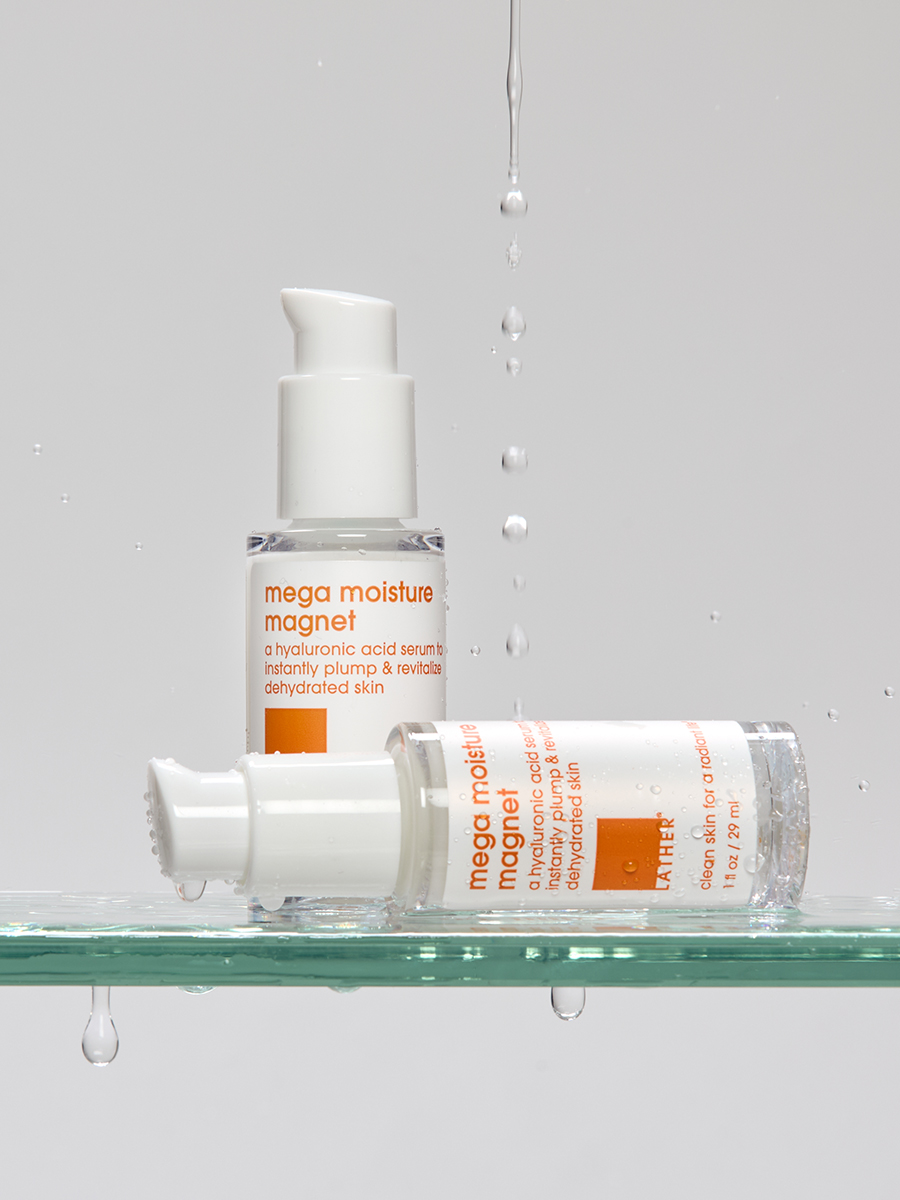 Two glass bottles of Lather mega moisture magnet hyaluronic serum tipped on a wet glass shelf beneath falling water droplets.
