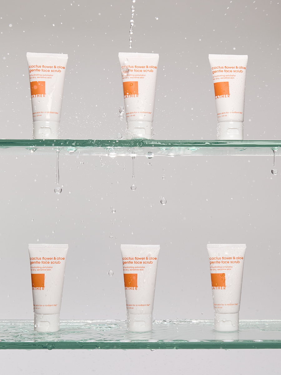Six tubes of Lather cactus flower & aloe face scrub arranged on two wet glass shelves as water rains down.