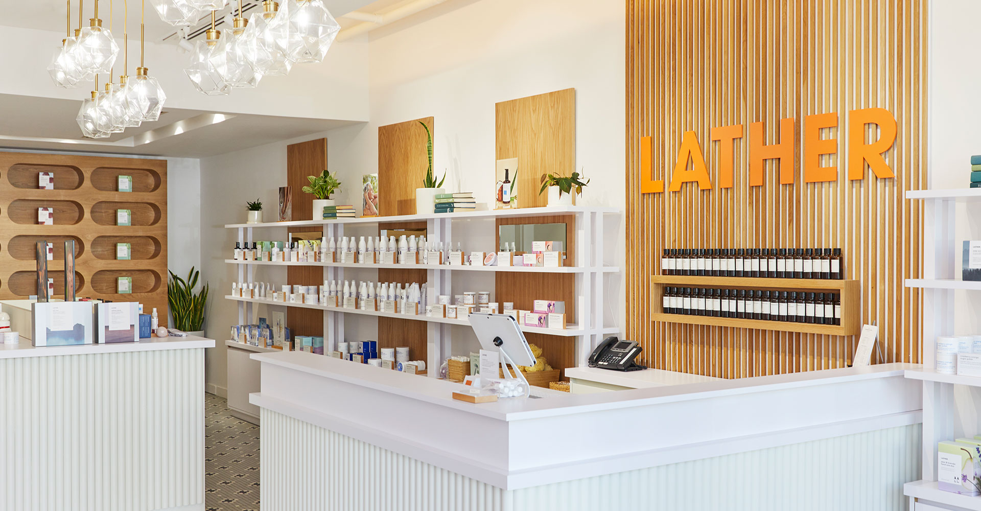 Lather Pasadena's front counter, with dimensional orange 'LATHER' wall lettering, oak apothecary shelving stocked with essential-oil bottles, glass pendant chandeliers, and a ridged white checkout counter on patterned tile flooring.