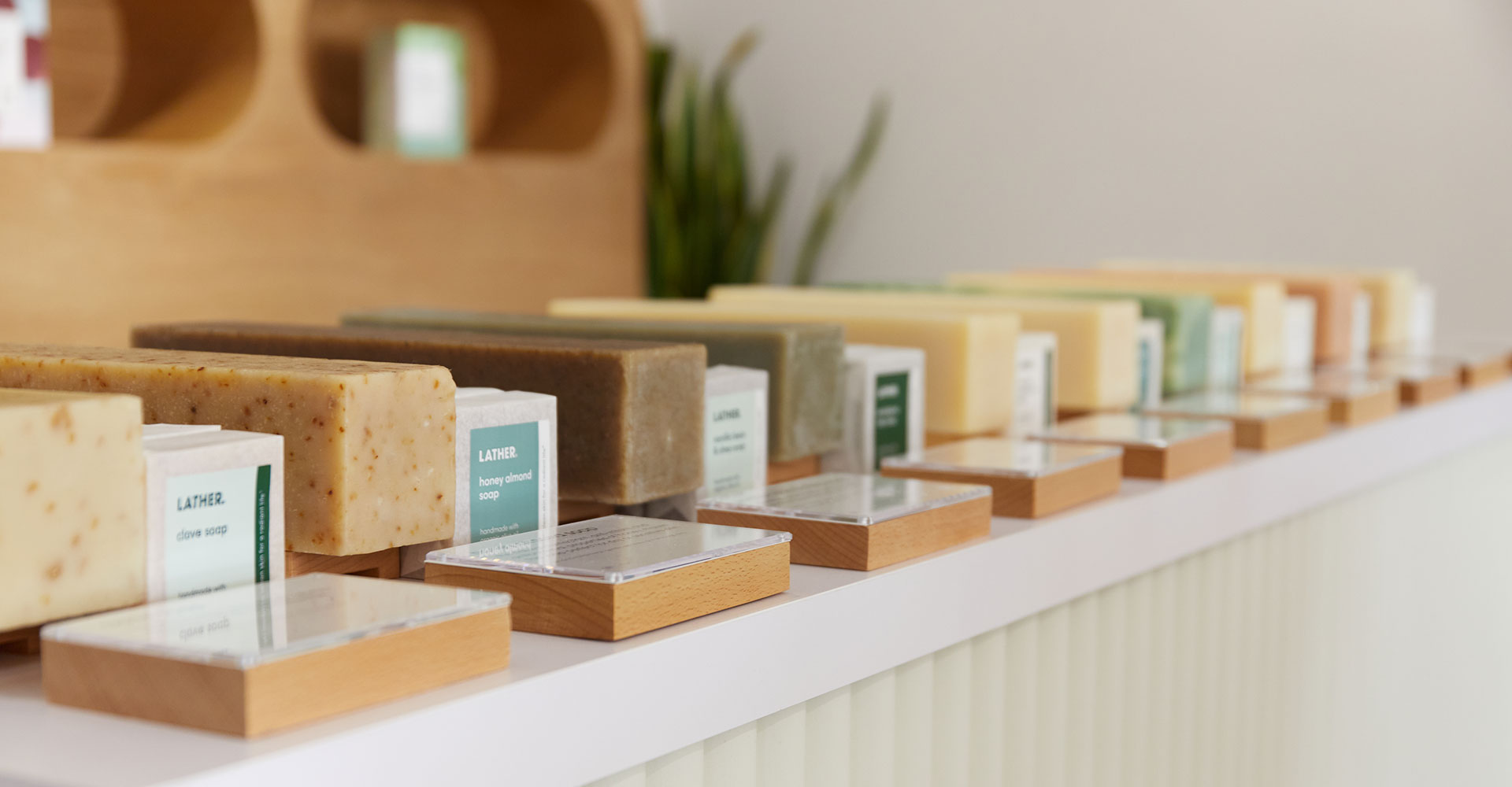 Handmade bar soaps in varied natural colors lined up on a white ridged shelf, each paired with a small 'Lather' label card in a wood stand.