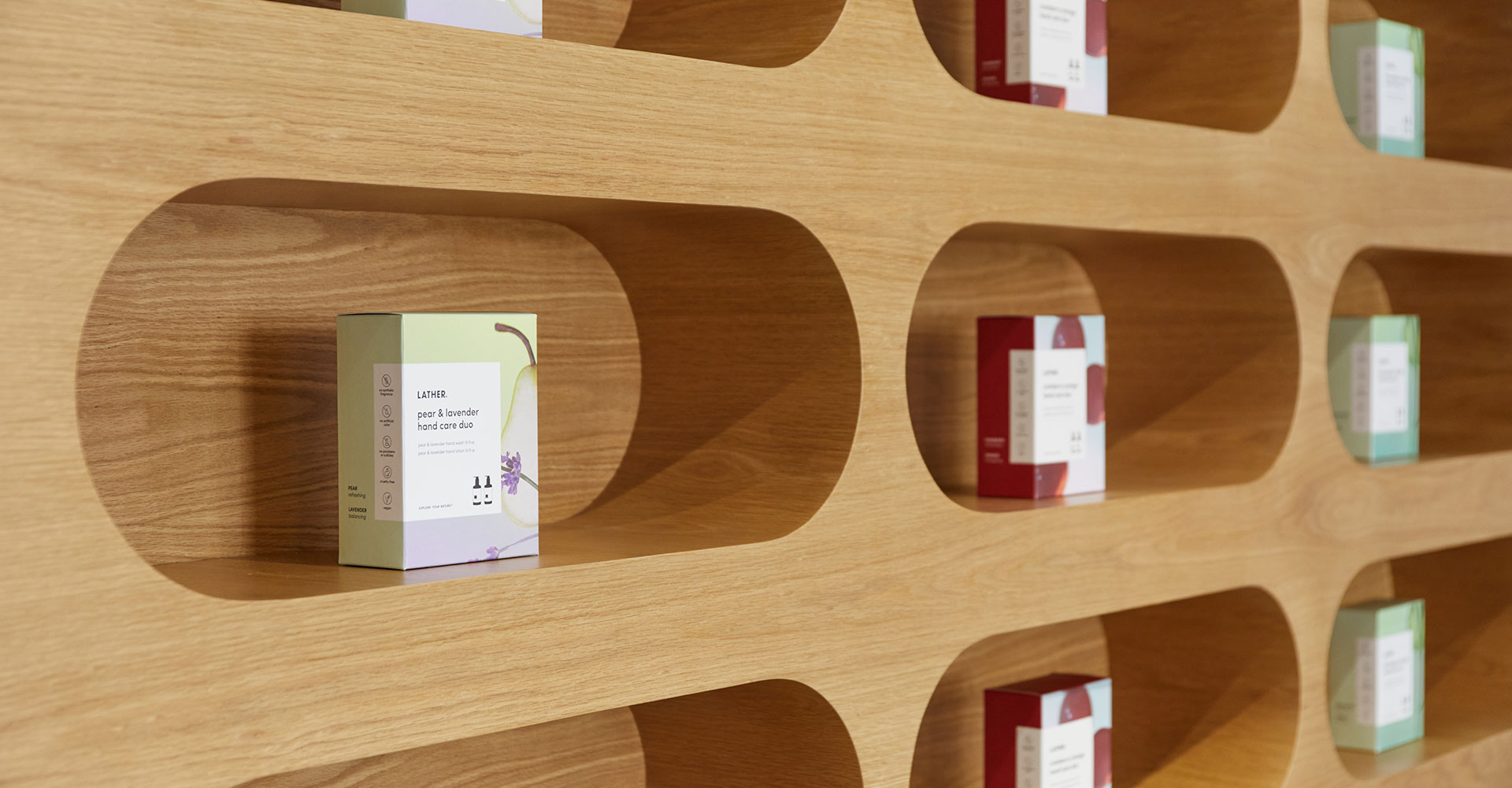 A boxed Lather 'Pear & Lavender Hand Care Duo' set inside one of the oak wall's rounded display niches, with red and green boxed products visible in the surrounding niches.