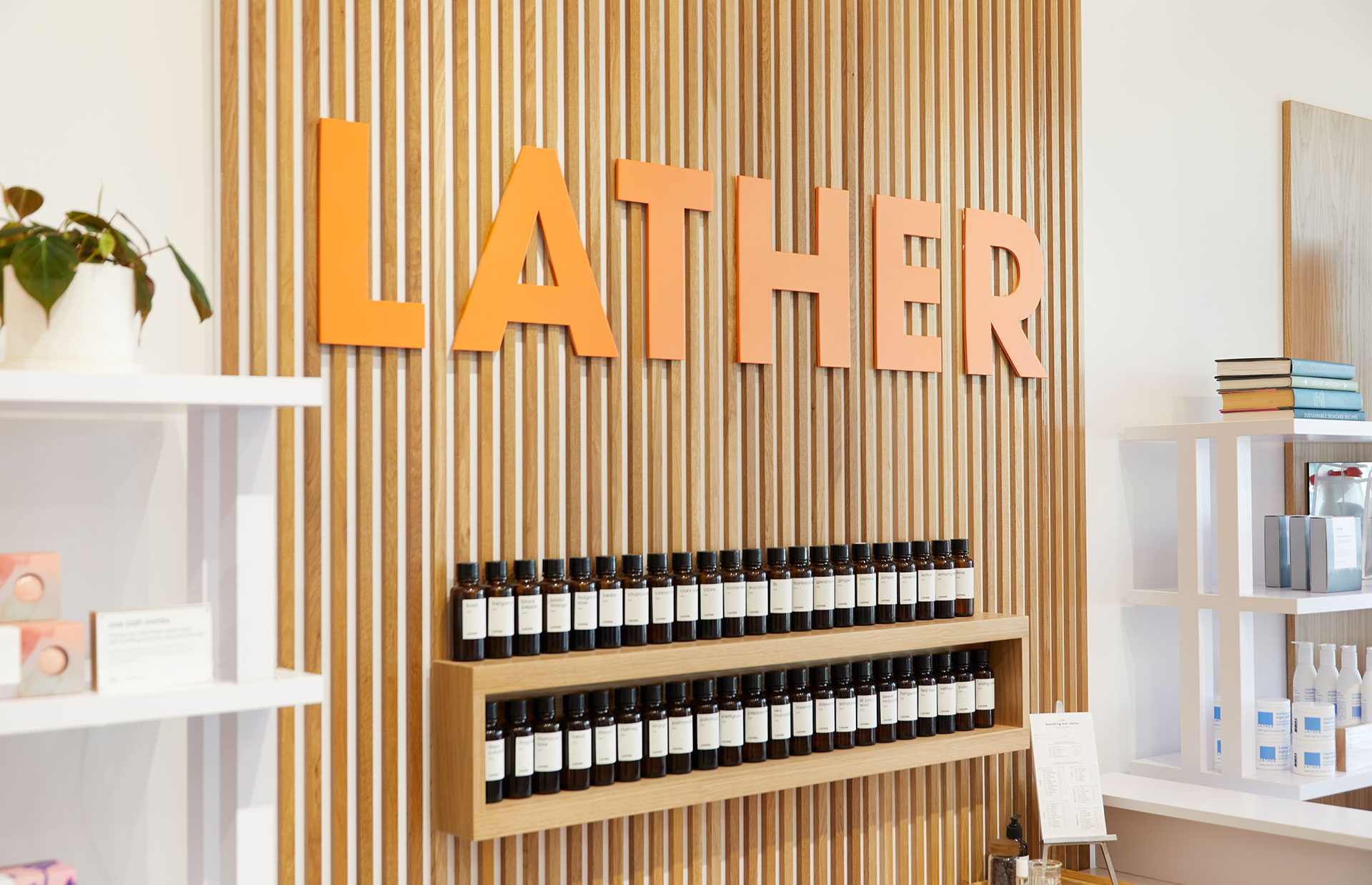The dimensional orange 'LATHER' wordmark mounted on a vertical oak-slat wall above two shelves of amber fragrance bottles, with white display shelving and a potted plant alongside.