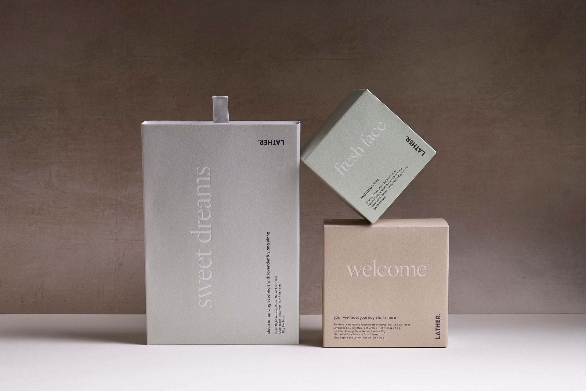 Three LATHER hotel amenity boxes styled together against a warm brown backdrop: a tall silver 'sweet dreams' sleep-essentials box, a sage-green 'fresh face' hydration trio box balanced on its corner, and a tan 'welcome' wellness box in front.