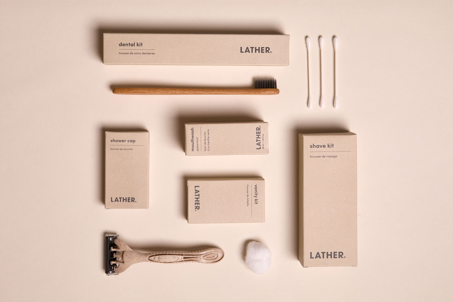 Flat-lay of LATHER travel amenity items on a cream ground: kraft boxes for a dental kit, shower cap, mouthwash, vanity kit, and shave kit, alongside a bamboo toothbrush, cotton swabs, a razor, and a cotton ball.