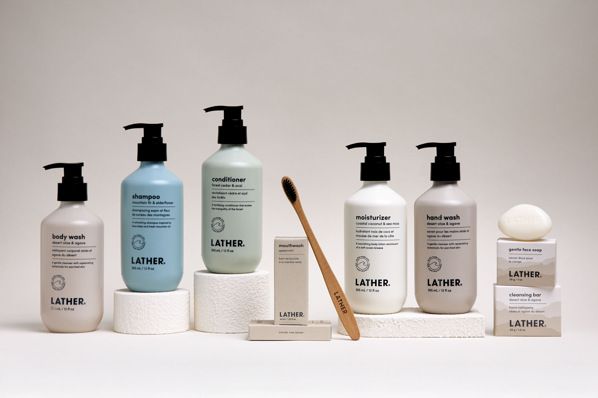 Full LATHER hotel bath collection lined up on a plain ground: body wash, shampoo, conditioner, moisturizer, and hand wash bottles with black pump caps, plus a boxed toothbrush, mouthwash, bar soap, and cleansing bar.