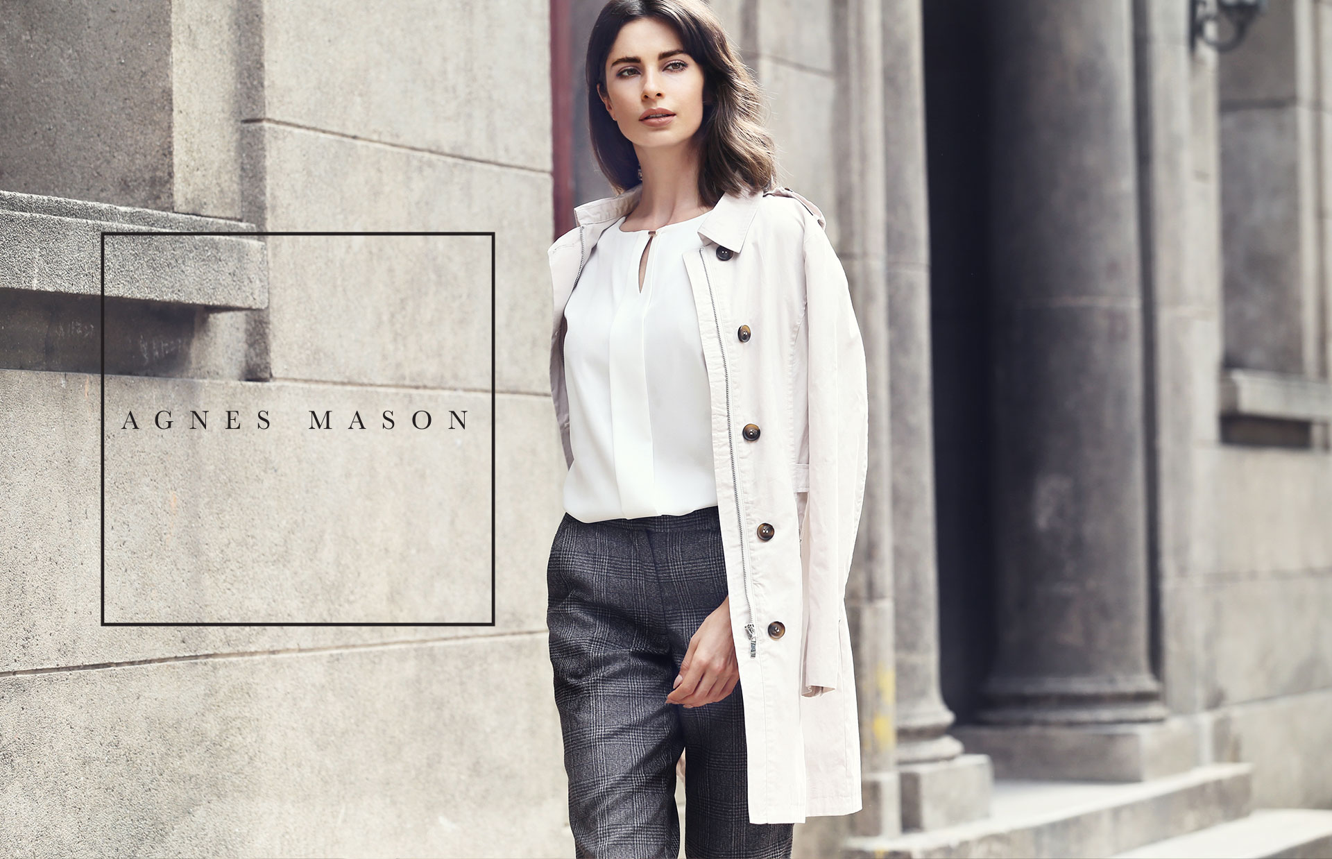 Agnes Mason logo — a letter-spaced wordmark inside a thin-lined square — set on a concrete wall beside a model in a tailored trench coat.